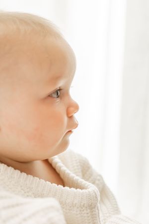 A baby wearing a white knit sweater gazes softly toward a window in gentle natural light.
