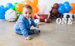 Baby in blue outfit sitting on floor with sports-themed birthday decorations and balloons.