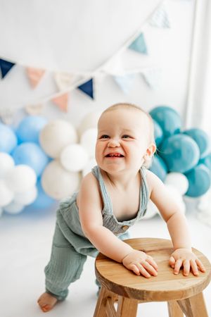 A baby in blue overalls leans against a wooden stool with birthday party decorations including balloons and banners behind.