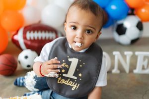 Baby wearing bib marked 'ONE' surrounded by sports balls and colorful balloons.