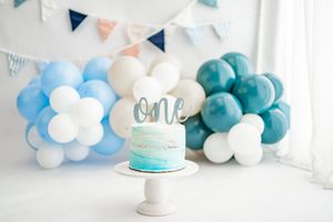 A turquoise smash cake sits on a white stand with blue and white balloon decorations and bunting for a first birthday.