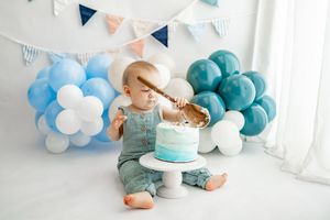 First birthday cake smash photo shoot with blue balloon arch and bunting decorations in white studio setting.