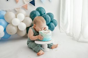 Small child wearing green outfit celebrates first birthday with smash cake photoshoot in blue and white decor.