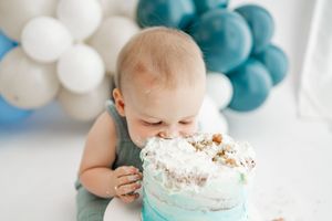 Baby smashing into white and blue birthday cake during cake smash photoshoot with teal and white balloons in background.