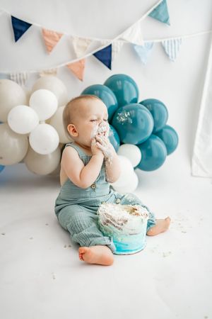 Baby in turquoise overalls enjoying cake during first birthday photo shoot with balloons and bunting decorations.