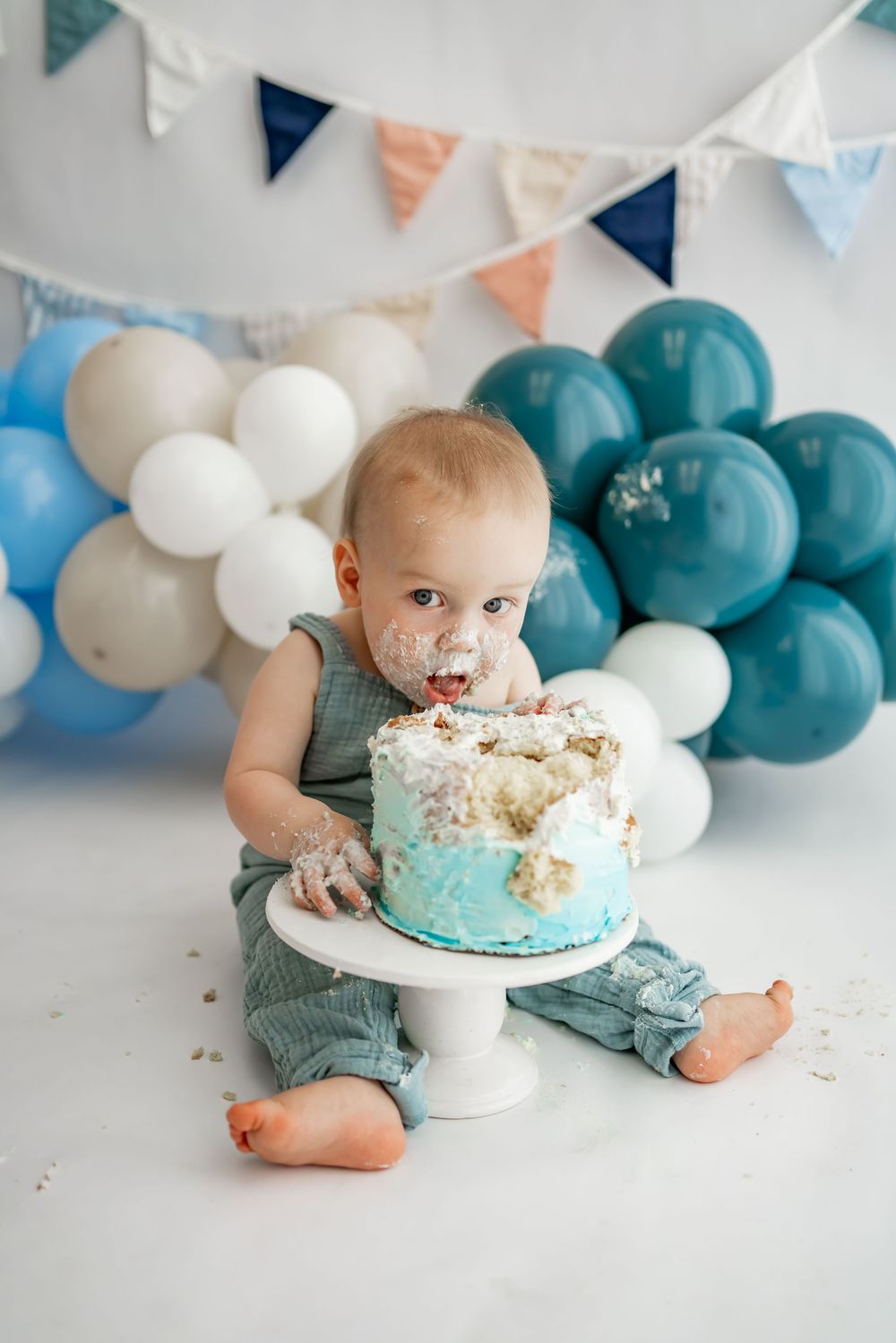 A smash cake photo session featuring teal and white balloons with a one year old enjoying birthday cake.