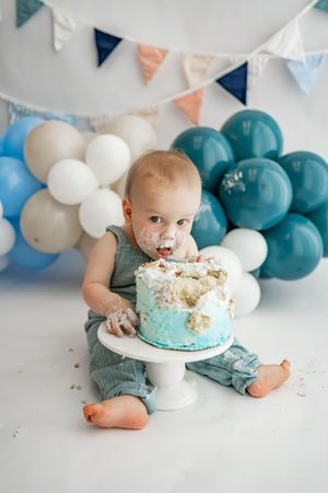A smash cake photo session featuring teal and white balloons with a one year old enjoying birthday cake.