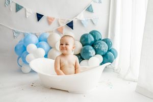 A baby sits in a white bathtub surrounded by blue and white balloons with a bunting banner overhead.