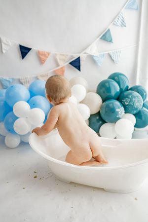 Baby sitting in white bathtub with blue and white balloon decorations and bunting flags in background.