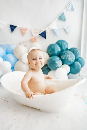 Baby sits in curved white basin surrounded by white and teal balloons with decorative bunting in background.