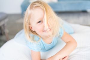 Child with blonde hair lying on white bedding wearing light blue dress.
