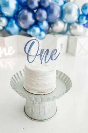Small white frosted cake displayed on vintage-style metal stand with blue and white balloon decorations in background.