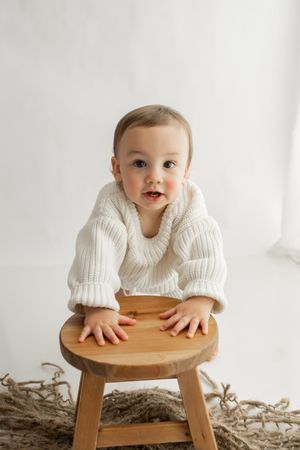 Baby in white sweater peeks over wooden stool in minimalist portrait session.