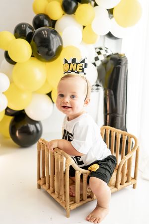 Baby sits in wooden crib surrounded by festive yellow, black and white balloons at party celebration.