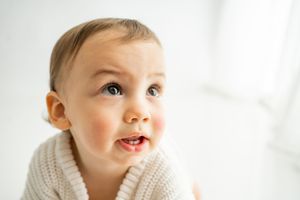 Toddler smiling and looking upward wearing a cozy white knit sweater in a bright setting.