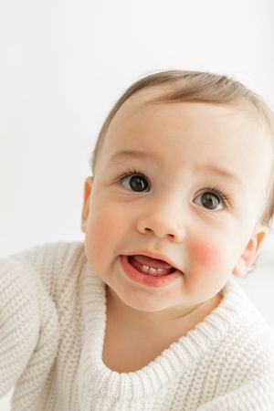 Close-up portrait of a baby wearing a white knit sweater with a bright smile and rosy cheeks.