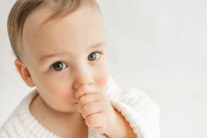 A small baby in white clothing lies on white bedding while touching their mouth and looking at the camera.