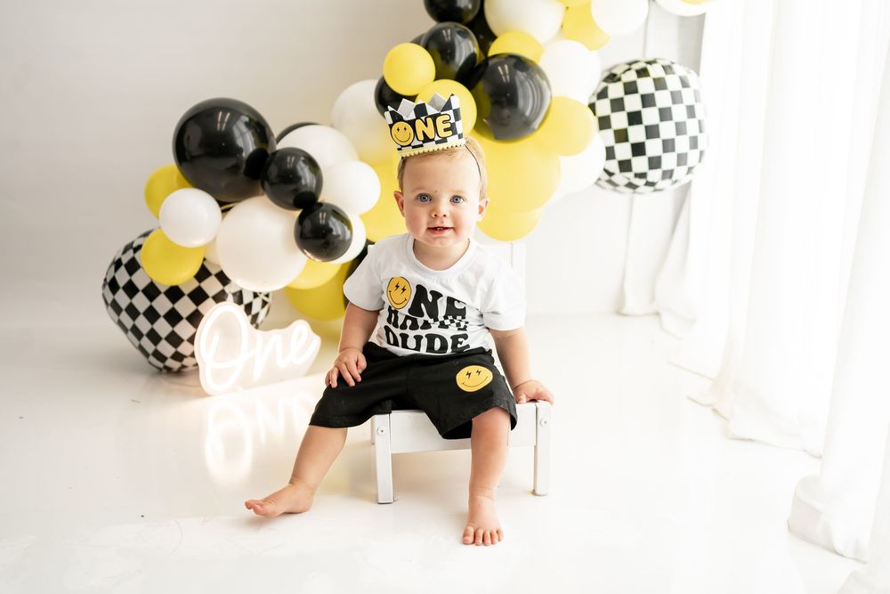 First birthday party setup with black and yellow balloons and checkered decorations on white backdrop.