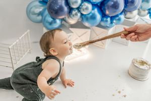 Baby in olive green overalls explores cake smash setup with blue balloons and white decorations during first birthday photoshoot.
