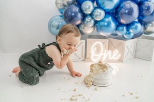Baby crawls towards birthday cake during cake smash photo session with blue balloon arch and white decorations.