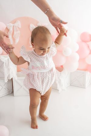 Baby in floral dress takes first steps with helping hands on white floor with pink balloons.