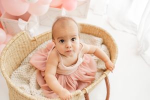 A small baby in a pink dress lies on a white blanket next to a woven basket, creating a sweet portrait setting.