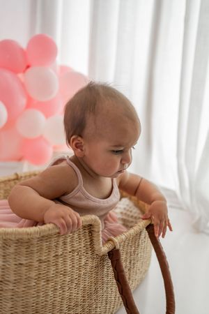 Baby explores woven basket from four different angles with pink heart balloons in background.