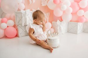 Baby in white dress sits on floor with pink and silver balloon decorations for first birthday celebration.