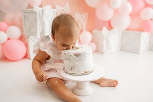 Small baby in pink and white dress holding pink balloons celebrates first birthday with pink and white decorations.