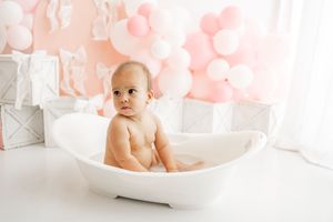 Baby with pink and white balloons celebrating first birthday party in bright festive room setting.