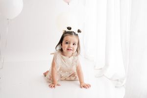 A smiling baby in white sits among balloons and flowers for a one-year-old milestone photo session.