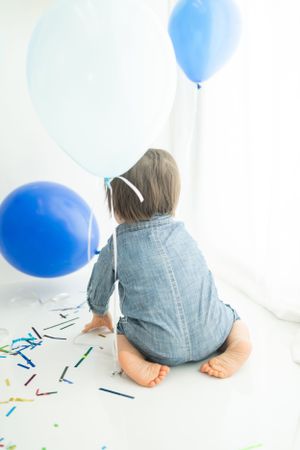 Back view of small child surrounded by blue balloons and colorful confetti on white floor.