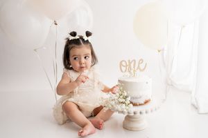 Baby wearing pink outfit sits on white floor next to elegant white birthday cake with floral decorations.