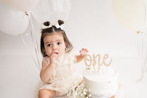 Baby wearing pink outfit sits on white floor next to elegant white birthday cake with floral decorations.