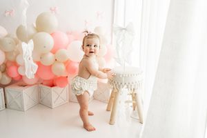 Baby in white outfit celebrating first birthday with pink and beige balloon decorations and floral accents.