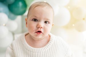 A little person in a white knit sweater sits with a bright teal background behind them.