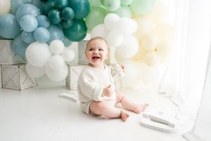 Baby in white onesie sits among teal and white balloon decorations for a birthday celebration.