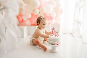 Baby in pink dress sits on white floor with birthday cake and decorations for first birthday celebration.