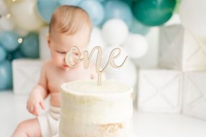 A white birthday cake with glowing 'one' topper sits in front of teal and white balloon decorations.