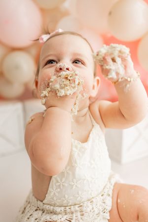 Baby in white lace dress with cake frosting on face smiles during first birthday cake smash photo session.