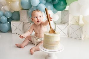 A white and sage green cake smash photo session capturing precious first birthday moments with balloons and decorations.
