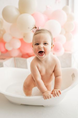 Joyful baby sits in white bathtub with pink and white balloons decorating background for birthday or celebration photo.
