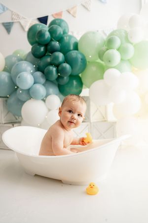 A bathtub photo session setup with teal and white balloon decorations and a rubber duck toy against a light backdrop.