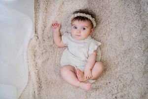 Baby wearing white outfit and headband lying on textured beige background.