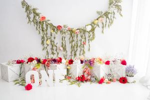 White decorative 'ONE' letters with silver presents and red roses arranged against a white backdrop with hanging greenery.
