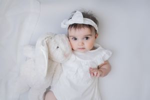 Baby in white dress lying on white fabric with plush bunny toy.