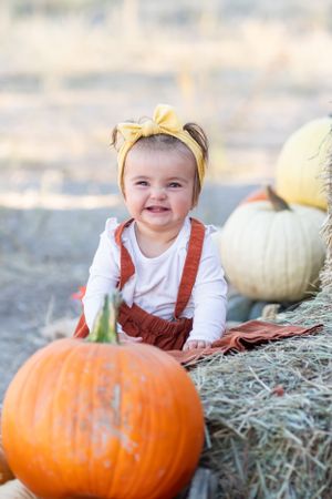 Baby wearing yellow headband and overalls poses with large pumpkin on hay bale.