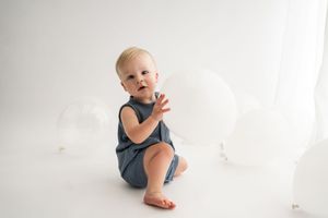 Baby wearing blue onesie sits and plays on white floor with natural backlighting.