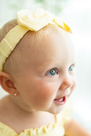 Adorable baby girl with blue eyes wearing a yellow headband with a flower and matching yellow outfit.