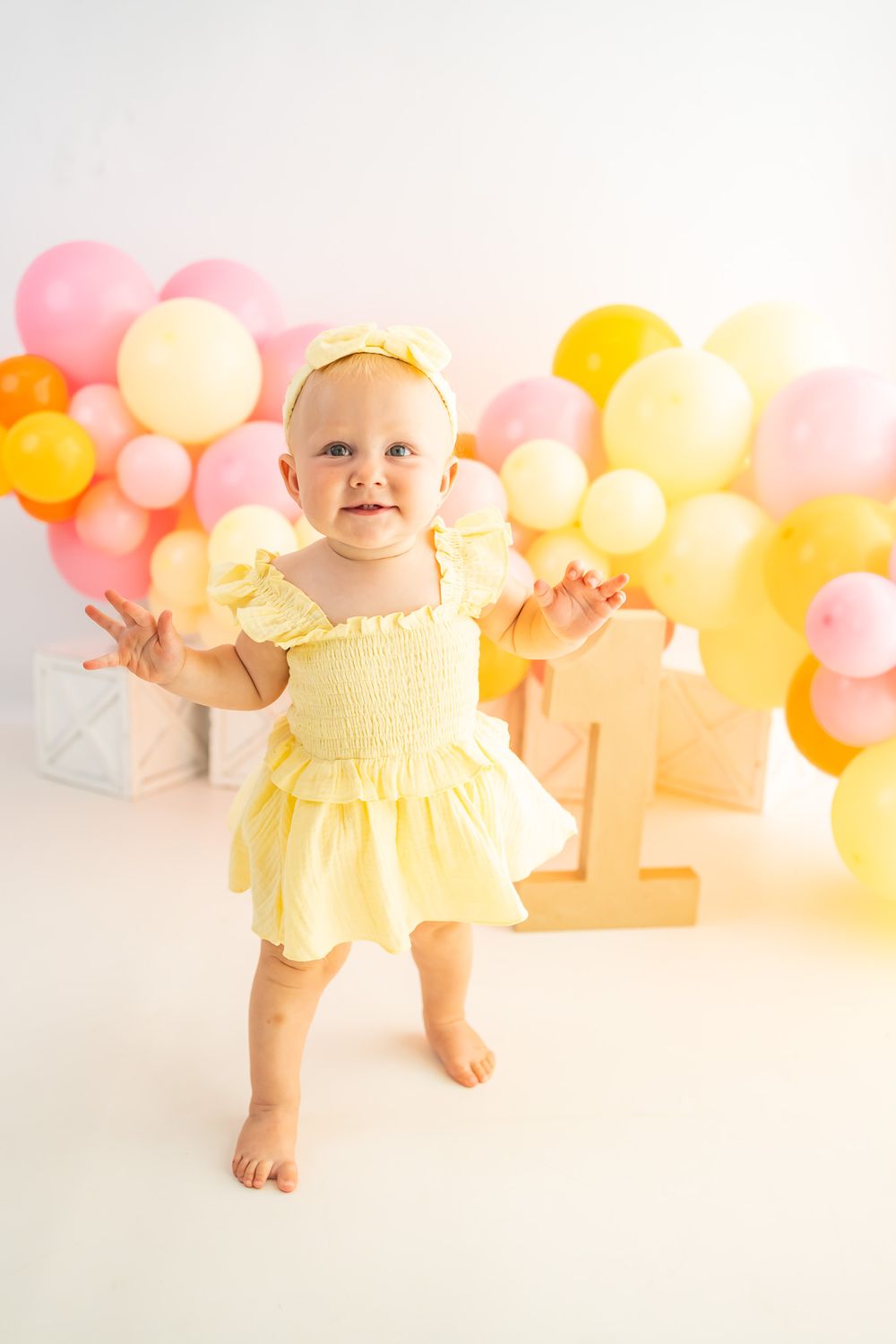 Adorable baby girl in yellow dress celebrating first birthday with colorful pink and yellow balloon backdrop.